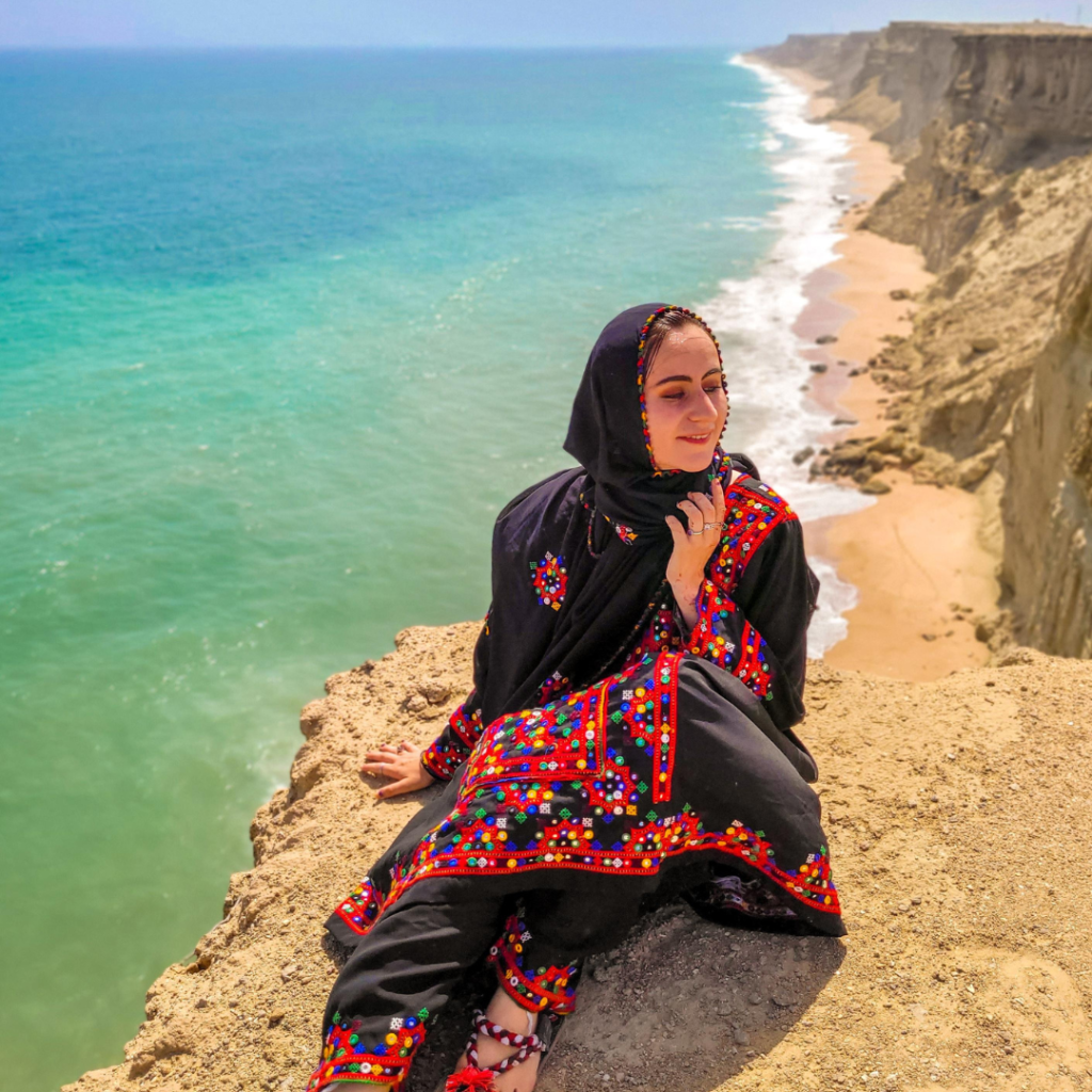 Girl in black traditional Balochi dress sitting on a cliff overlooking the turquoise Arabian Sea on the Makkoran Coast near Chabahar, Sistan and Baluchistan, Iran.