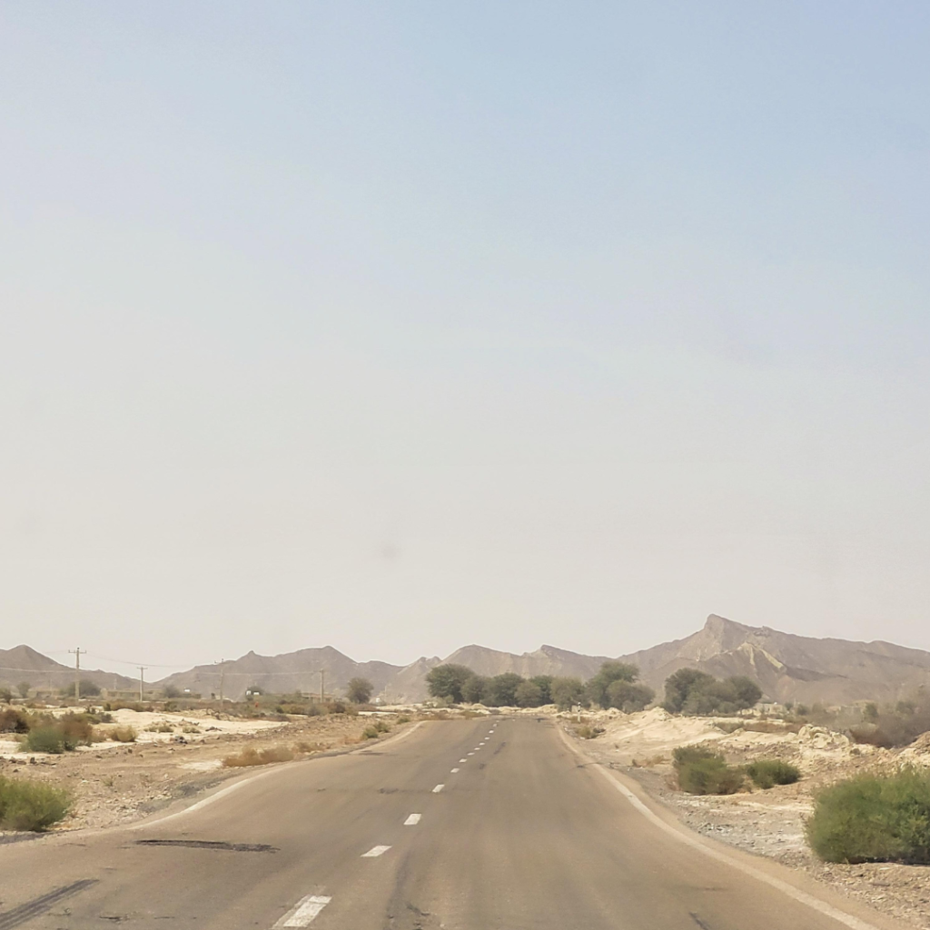 The road from the Iran-Pakistan border crossing at Rimdan towards Chabahar, Iran. An empty road leading through barren desert lands with green bushes and shrubs and dry mountains in the background.