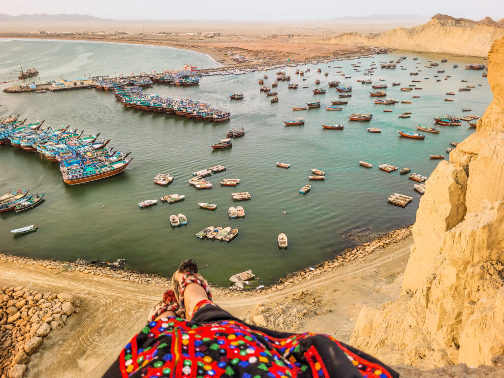 20 Stunning Places To Visit In Chabahar, Iran 21 Girl in traditional Balochi dress and shoes sitting at the edge of a cliff overlooking a turquoise bay with fishing boats in Beris, Iran.