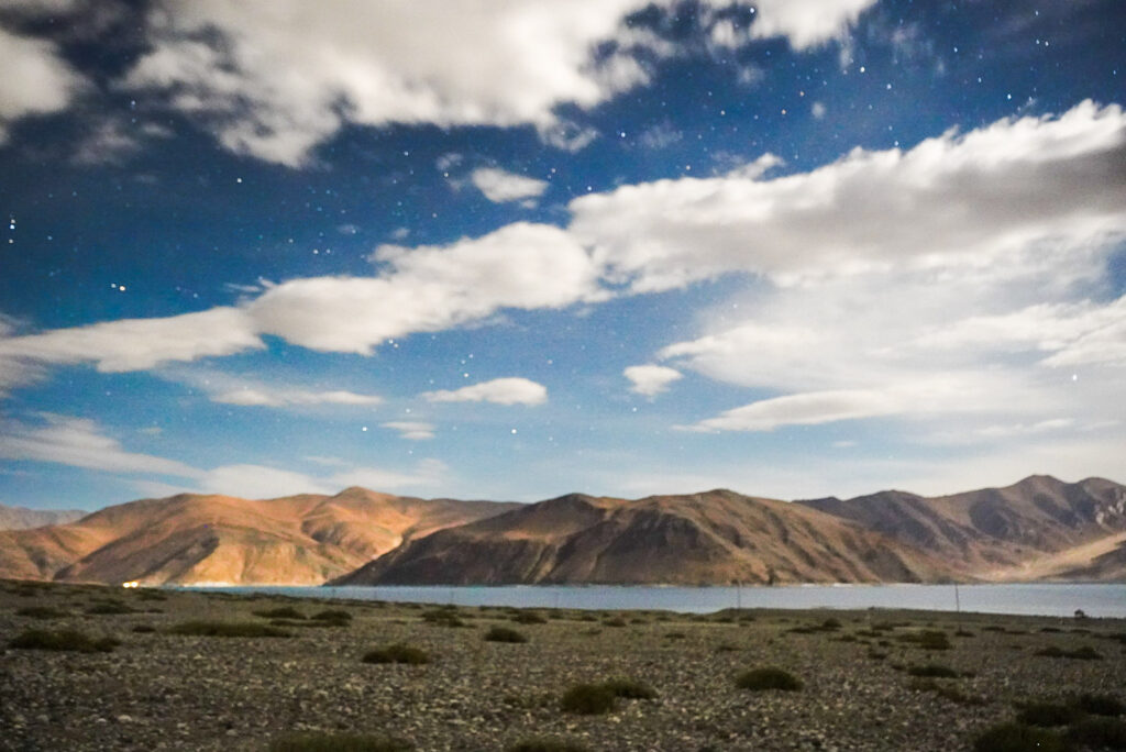 Pangong Tso Lake captured at night with white clouds and stars in the sky.