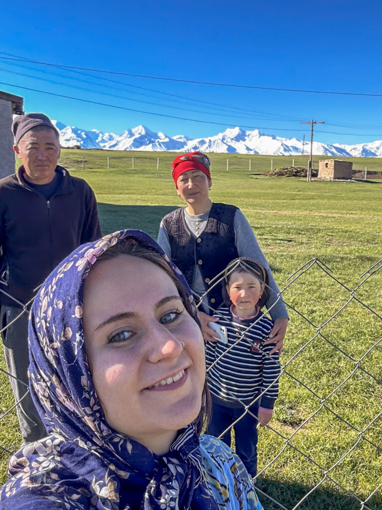 Tourist girl posing with a Kyrgyz family on a green meadow with white mountains in the background in Sary Tash, Kyrgyzstan