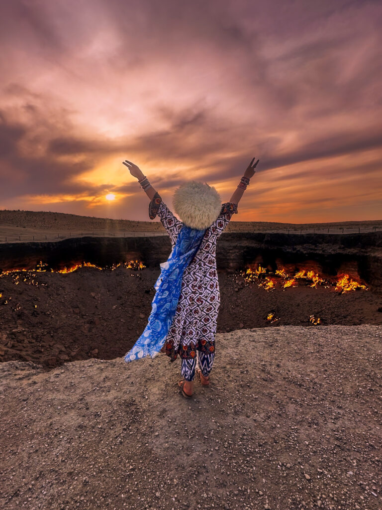 Tourist girl posing in front of the burning gas crater 'Derweze; Gate to Hell' in Turkmenistan during sunset, wearing a traditional Turkmen wool hat