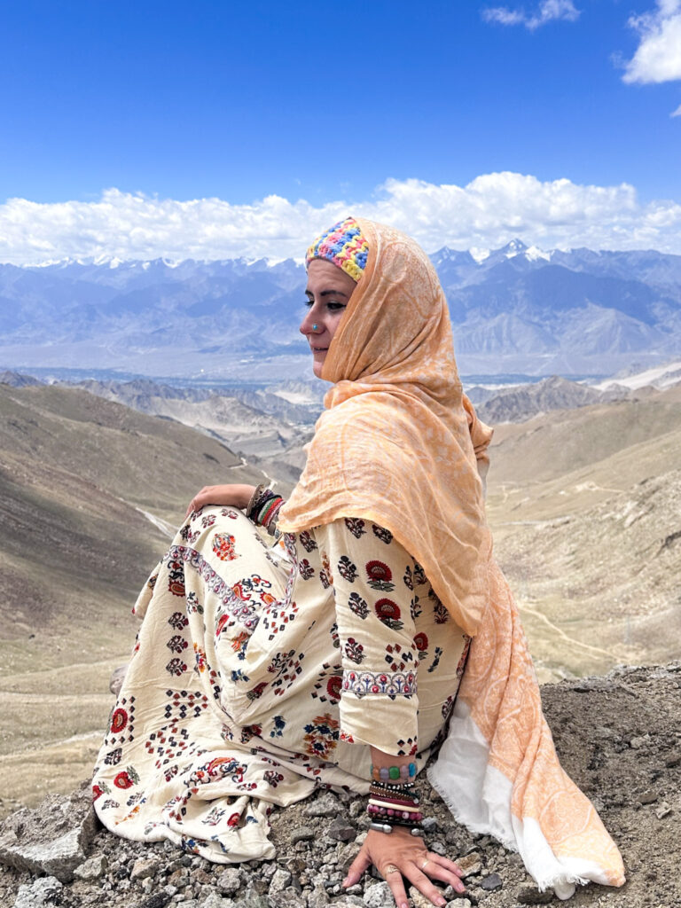 Foreign tourist girl in Indian dress and cream-colored Kashmiri veil sitting on the cliff of Khardung-La Pass, one of the world's highest motor able roads in Ladakh, India. There are barren valleys and snow-capped mountains in the background.